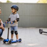 Five-year-old wearing a helmet on a three-wheel kick scooter while an electric scooter stays parked